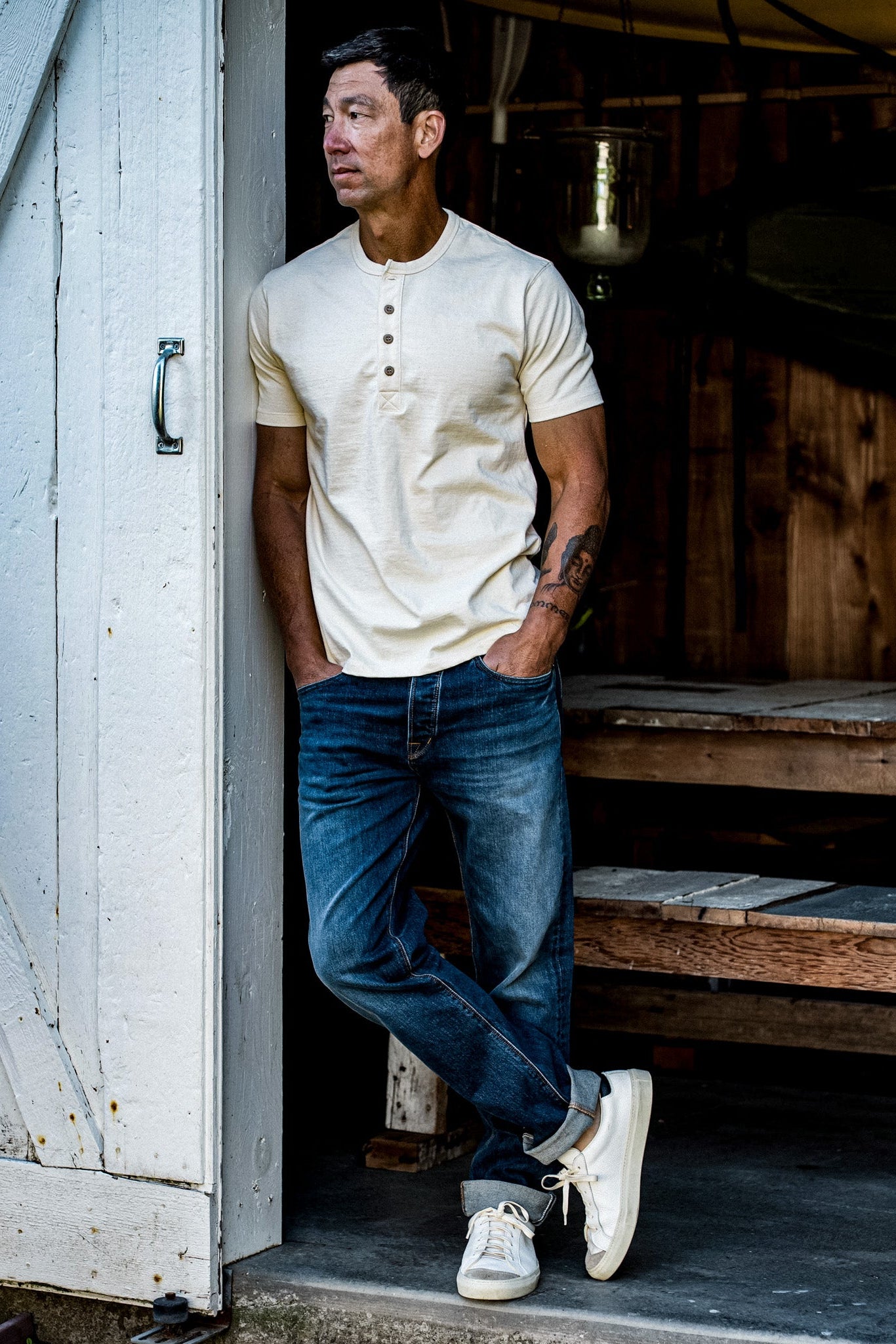 Man leaning in a barn doorway wearing a natural short-sleeve Bolinas Henley with cuffed selvedge denim jeans.