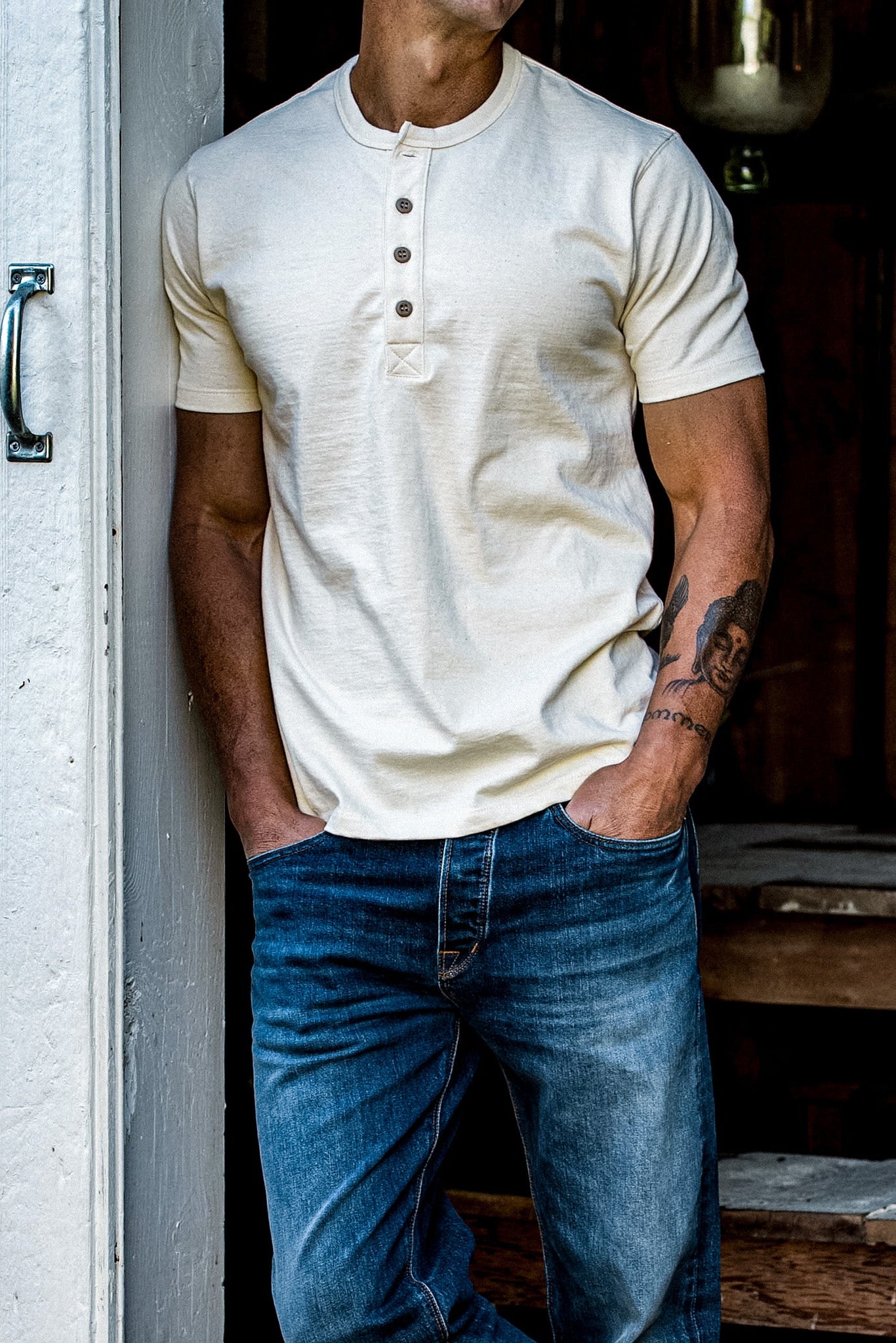 Man in a natural cream four-button Bolinas Henley and blue denim jeans leaning in a doorway.