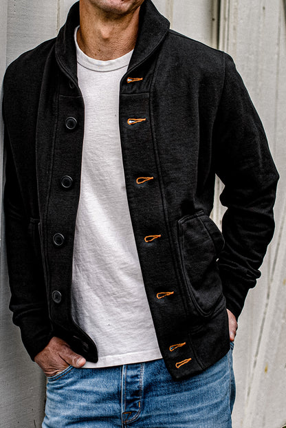 Man wearing a black shawl collar French terry cardigan with contrast orange keyhole stitching over a white tee and blue.