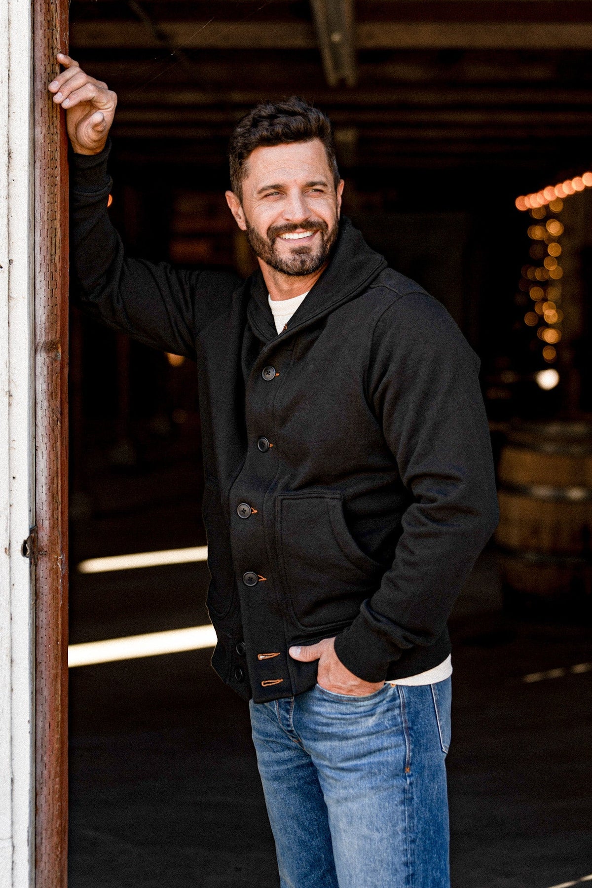 Smiling man leaning in a doorway wearing a men's basalt French terry cardigan over a cream tee and jeans