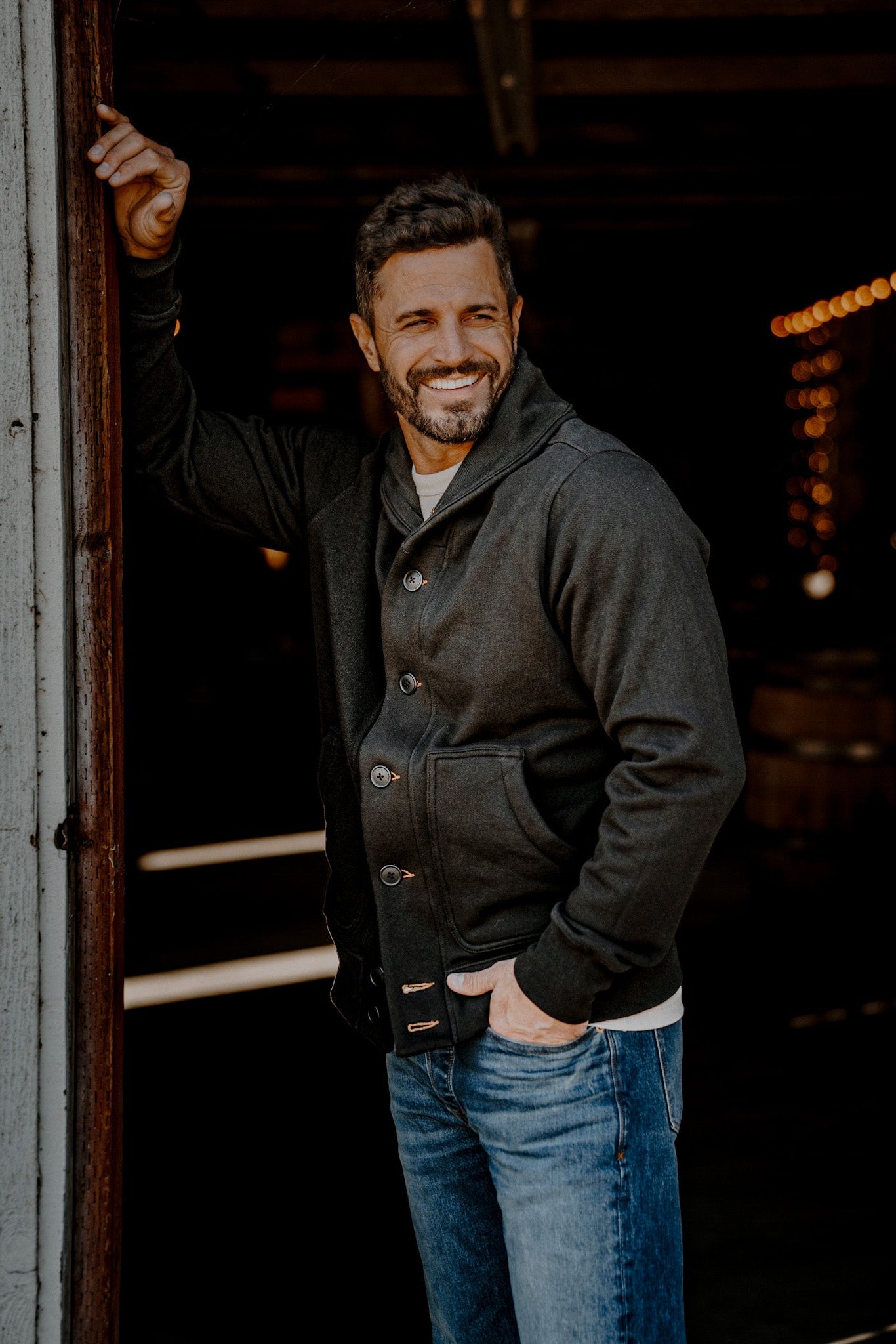 Laughing man leaning in a warehouse doorway wearing a men's basalt French terry cardigan and jeans
