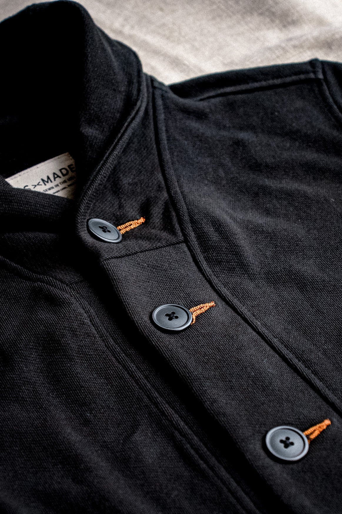 Close-up of the Bodega shawl cardigan in basalt black French terry with gray buttons and contrast tobacco-orange keyhole.