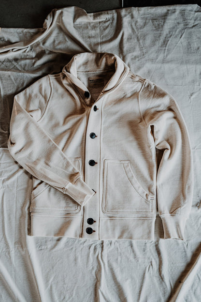 Flat lay of a natural cream men's shawl collar cardigan with dark buttons and patch pockets on a linen sheet.