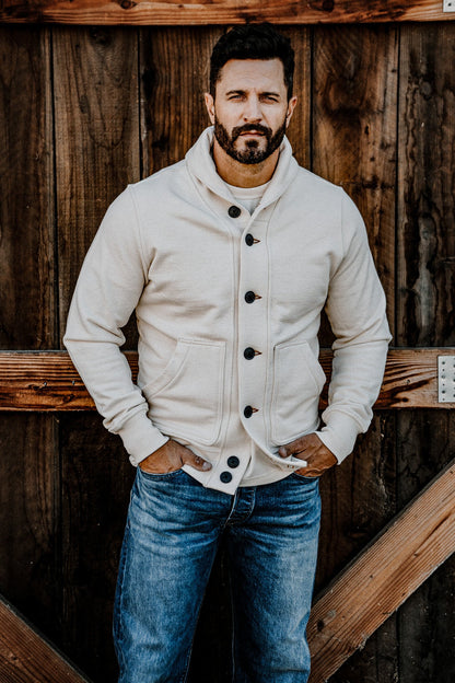 Man wearing a men's natural French terry shawl-collar cardigan over a henley and jeans, standing by a wood barn door
