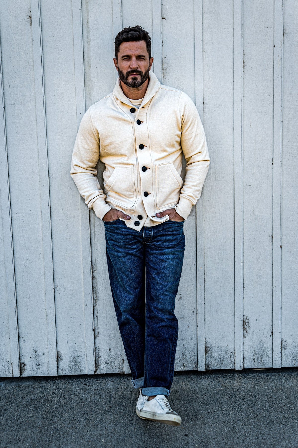 Man wearing a cream shawl collar cardigan, dark wash jeans, and white sneakers against a white barn door.