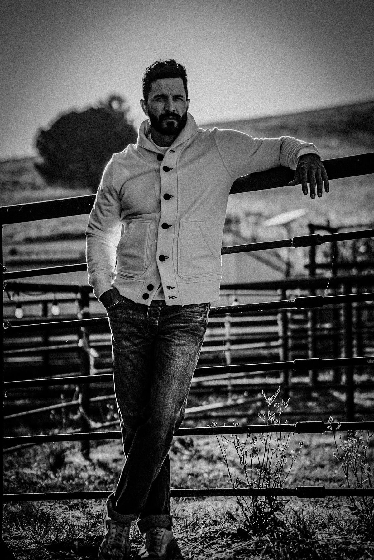 Black and white photo of a man in a shawl collar cardigan and jeans leaning on a metal ranch gate in an open field.