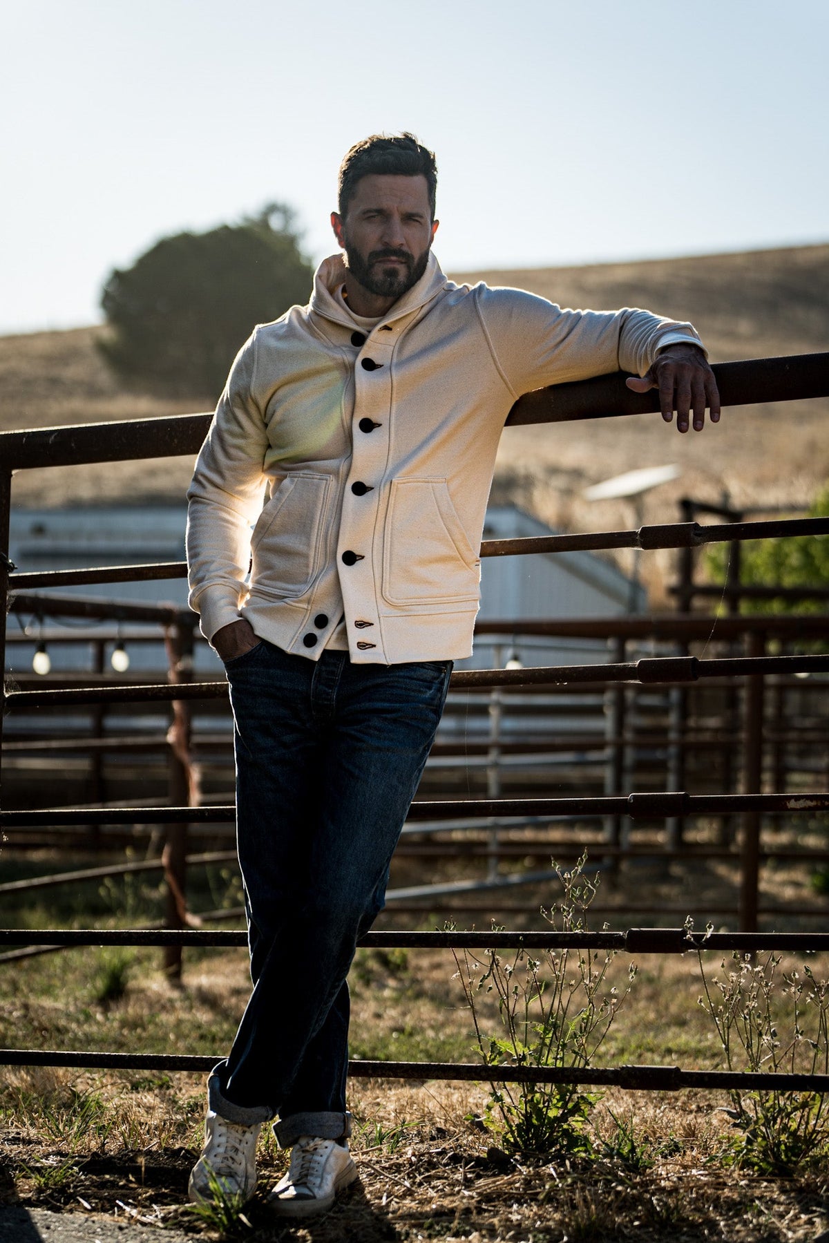 Man wearing a natural cream shawl collar cardigan and dark jeans leaning on a metal ranch gate at golden hour.