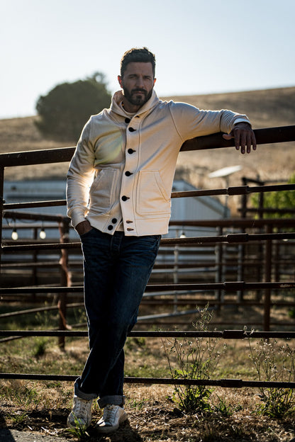 Man wearing a natural cream shawl collar cardigan and dark jeans leaning on a metal ranch gate at golden hour.