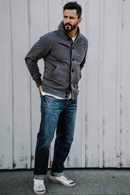 Man wearing a men's pebble gray French terry cardigan with jeans, standing against a wood plank wall
