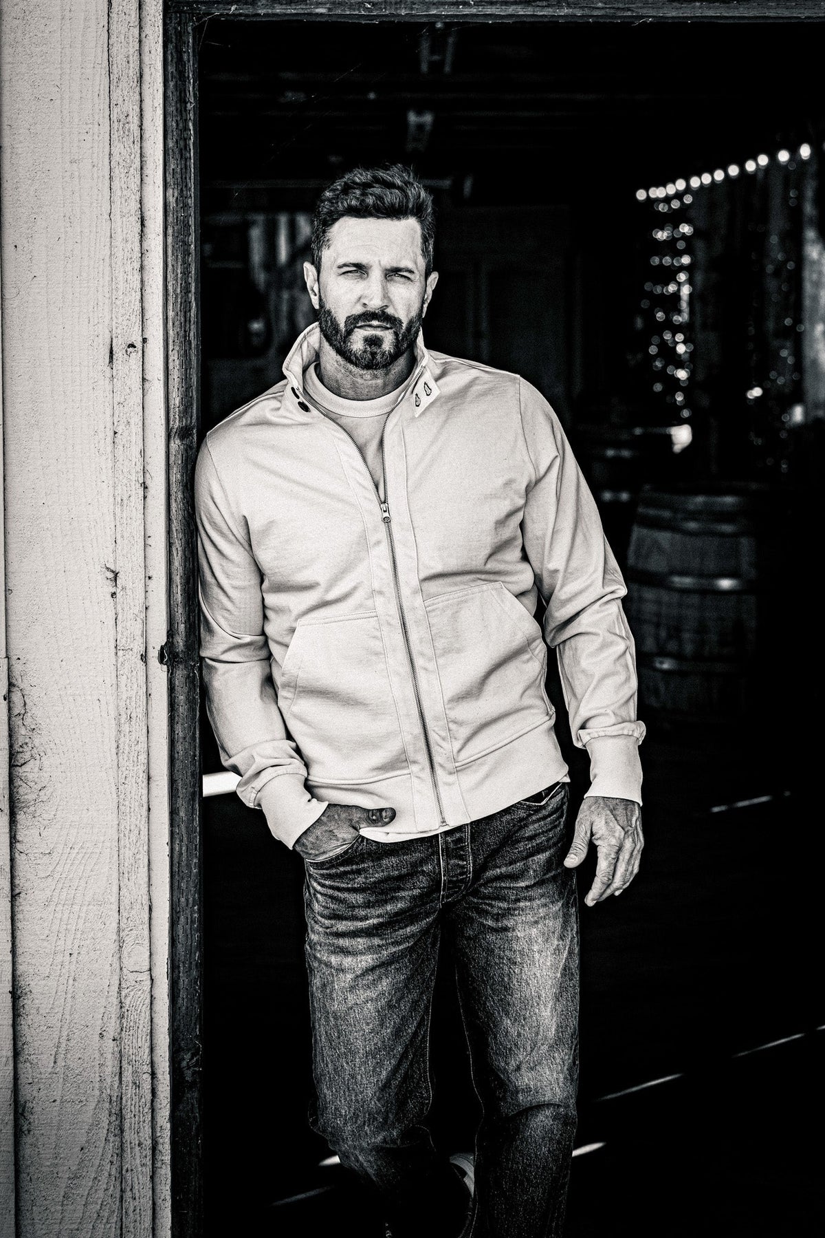 Black and white shot of a man in a men's zip-up moto jacket and jeans leaning on a barn door frame