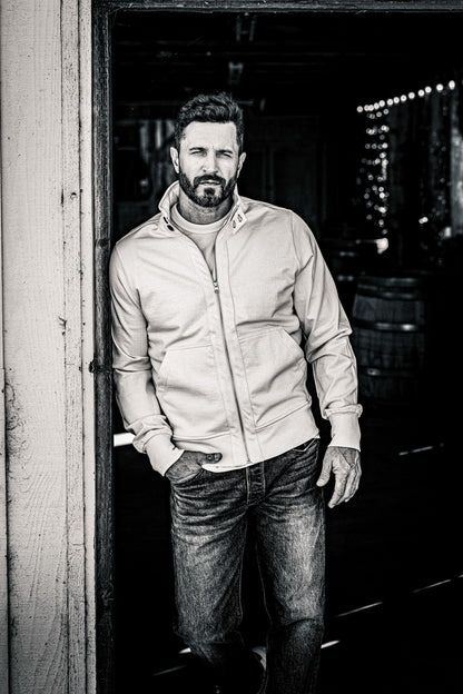 Black and white shot of a man in a men's zip-up moto jacket and jeans leaning on a barn door frame