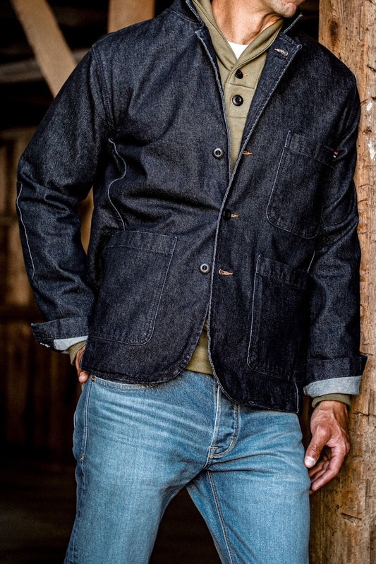 Men's denim blazer in dark indigo worn over an olive henley with light wash jeans, leaning against a wood beam.