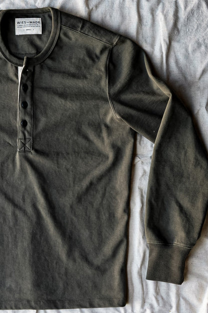 Flat lay detail of an olive cotton henley on linen with button placket and stitched neck label.