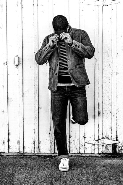 Black and white photo of a man in a men's ocean denim chore blazer buttoning the collar while leaning against a weathered wood barn door, one foot propped on the wall