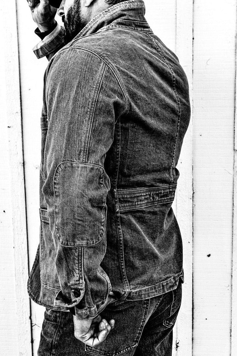 Black and white side-profile photo of a man in a men's wheat denim chore blazer and jeans against a wood wall