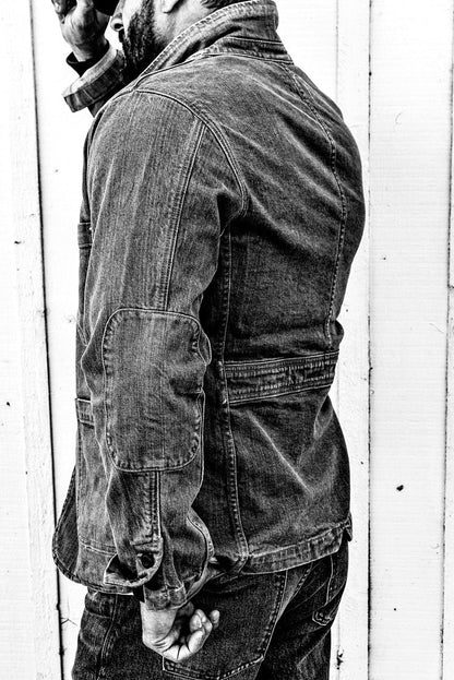 Black and white side-profile photo of a man in a men's wheat denim chore blazer and jeans against a wood wall