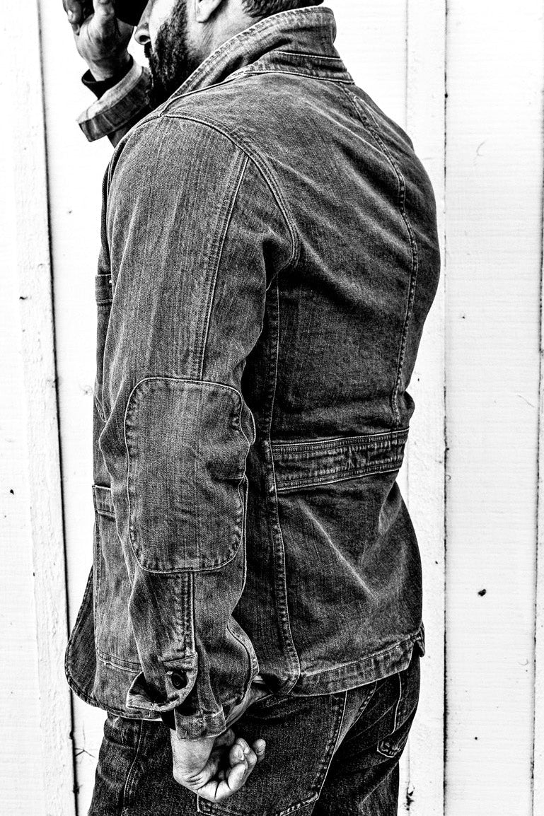 Black and white side-profile photo of a man wearing a men's ocean denim chore blazer and jeans against a weathered wood plank wall