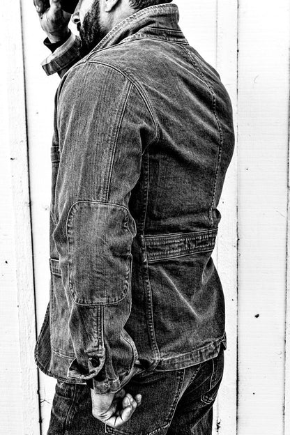 Black and white side-profile photo of a man wearing a men's ocean denim chore blazer and jeans against a weathered wood plank wall