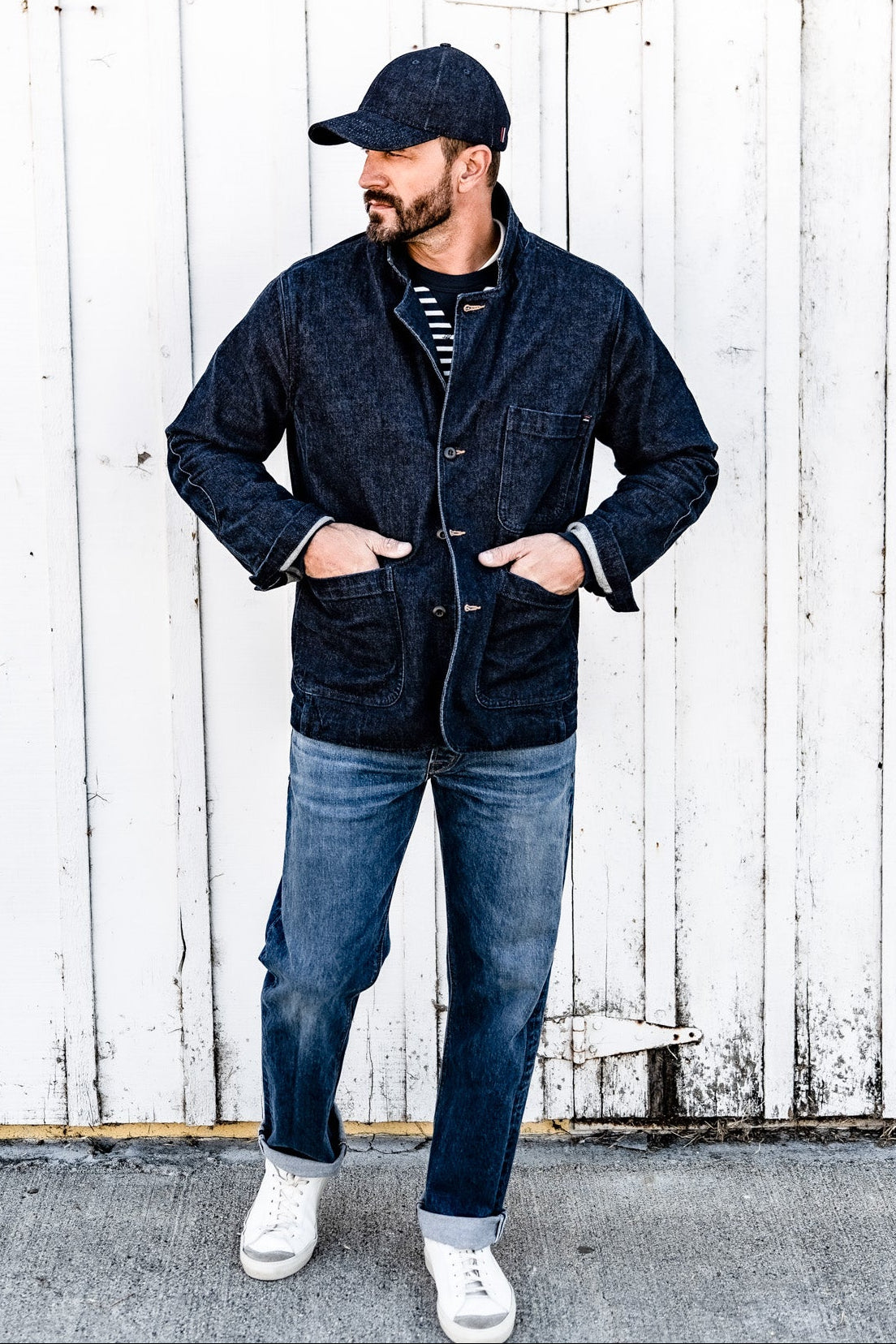 Full-body shot of a man in a cap wearing a men's ocean denim chore blazer, jeans, and white sneakers