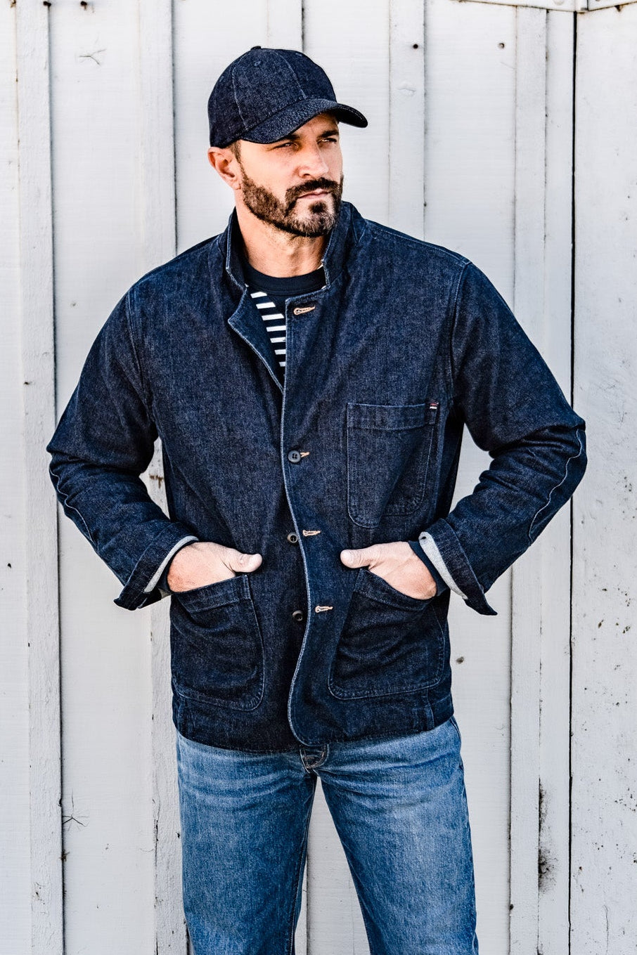 Close-up front view of a man in a cap wearing a men's ocean denim chore blazer with hands in the pockets