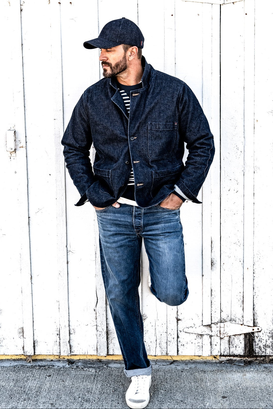 Man leaning against a wood plank wall wearing a men's ocean denim chore blazer, striped shirt, and jeans