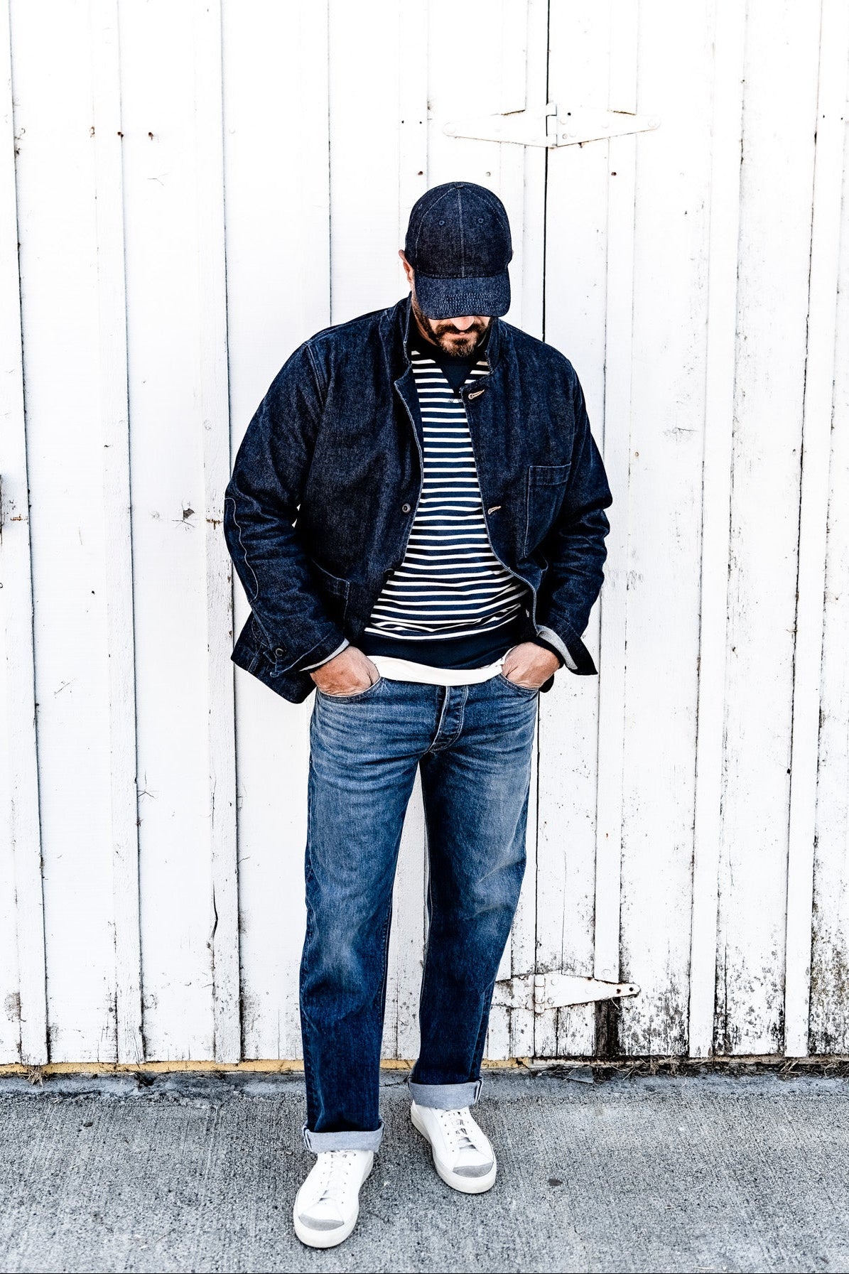 Man wearing a denim cap and a men's ocean denim chore blazer over a striped shirt with jeans, standing against a white wood plank wall
