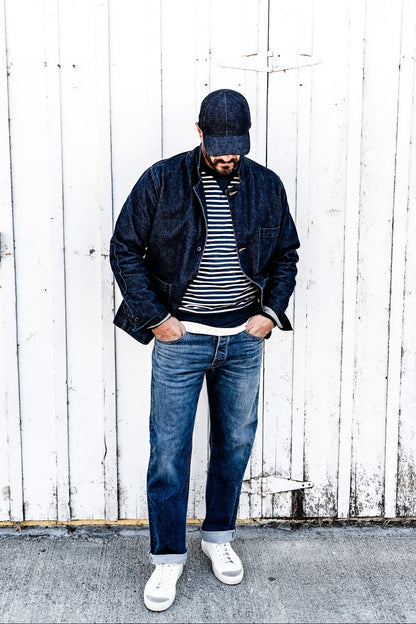 Man wearing a denim cap and a men's ocean denim chore blazer over a striped shirt with jeans, standing against a white wood plank wall