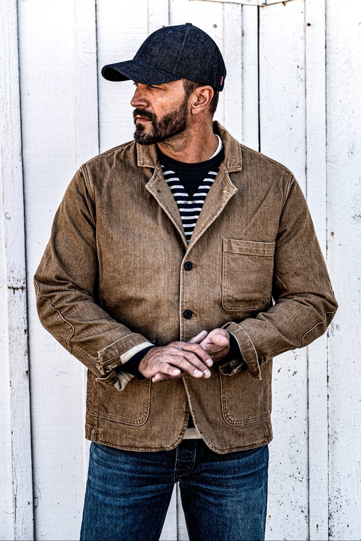 Man in a men's wheat denim chore blazer and jeans standing against a wood plank wall, hand in pocket