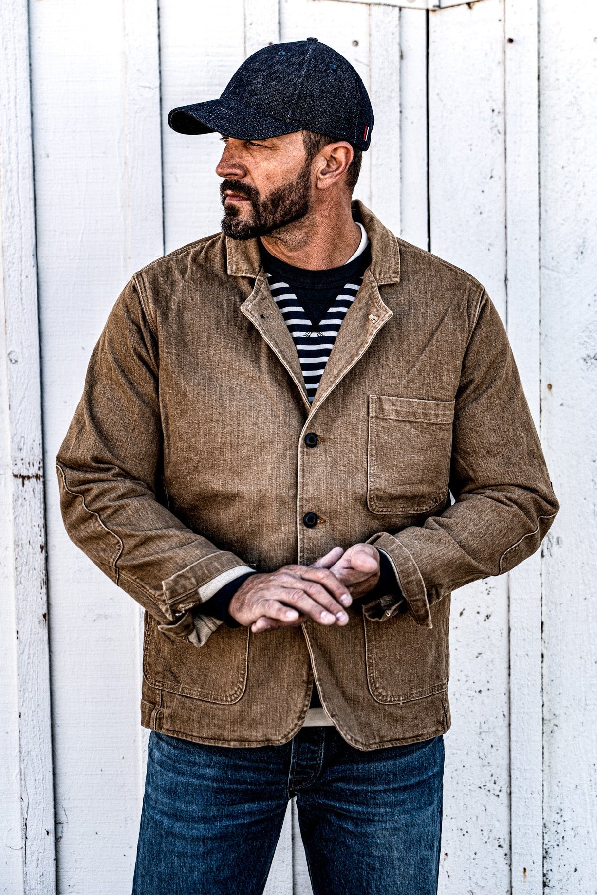 Man in a men's wheat denim chore blazer and jeans standing against a wood plank wall, hand in pocket