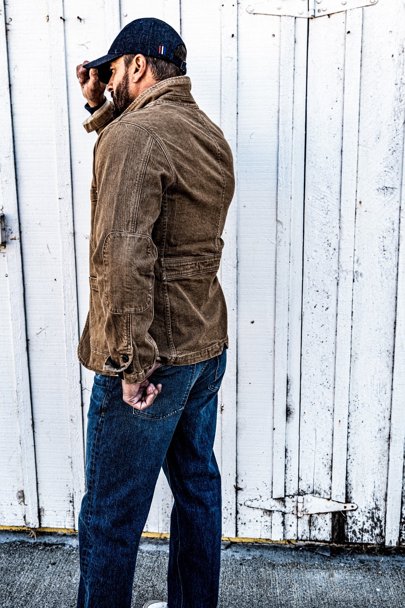 Side profile of a man in a men's wheat denim chore blazer and jeans, walking away from a wood plank wall
