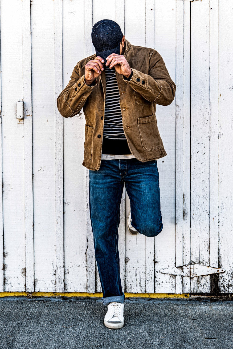 Man in a men's wheat denim chore blazer buttoning the collar while standing on one leg against a white barn door, wearing cuffed jeans and white sneakers