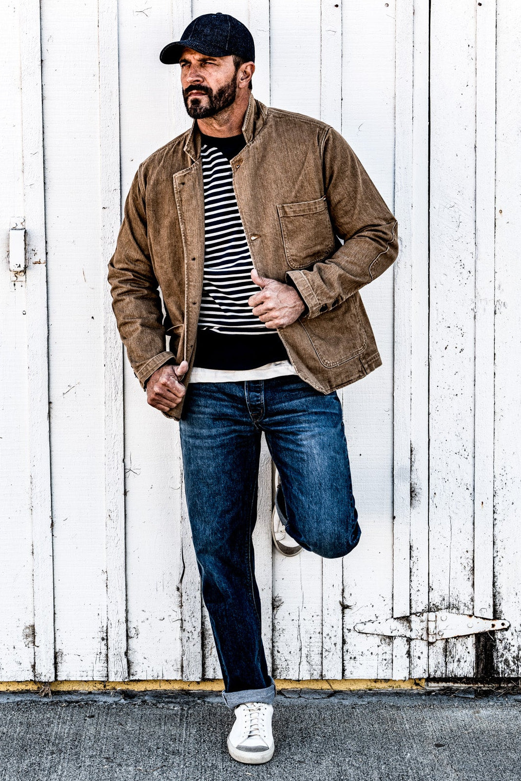 Man in a men's wheat denim chore blazer and jeans standing with one leg raised against a wood plank wall