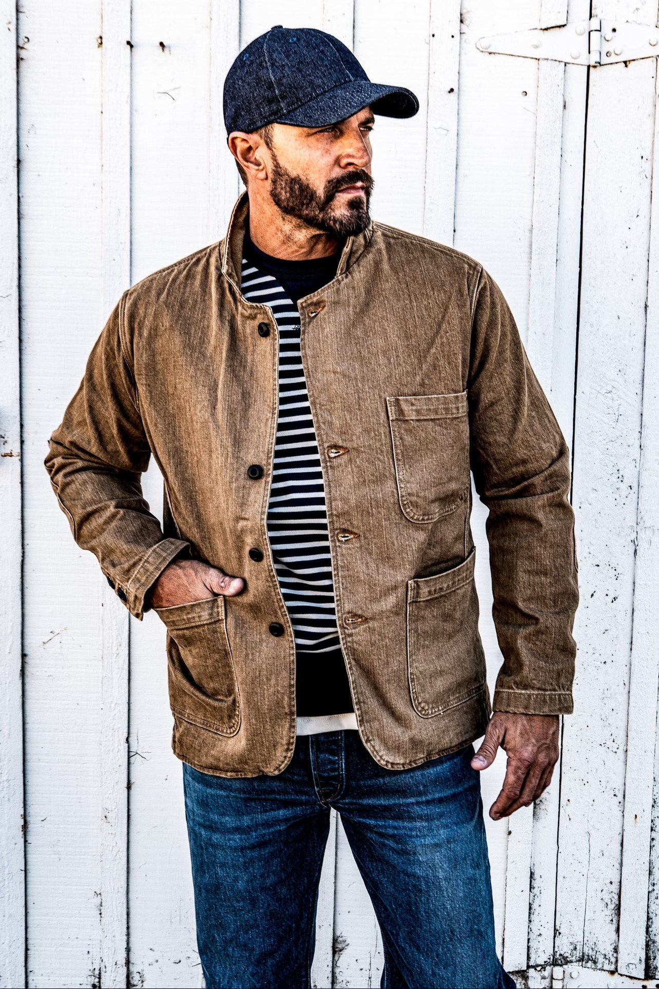 Close-up of a man wearing a men's wheat denim chore blazer open over a striped shirt, hand in pocket