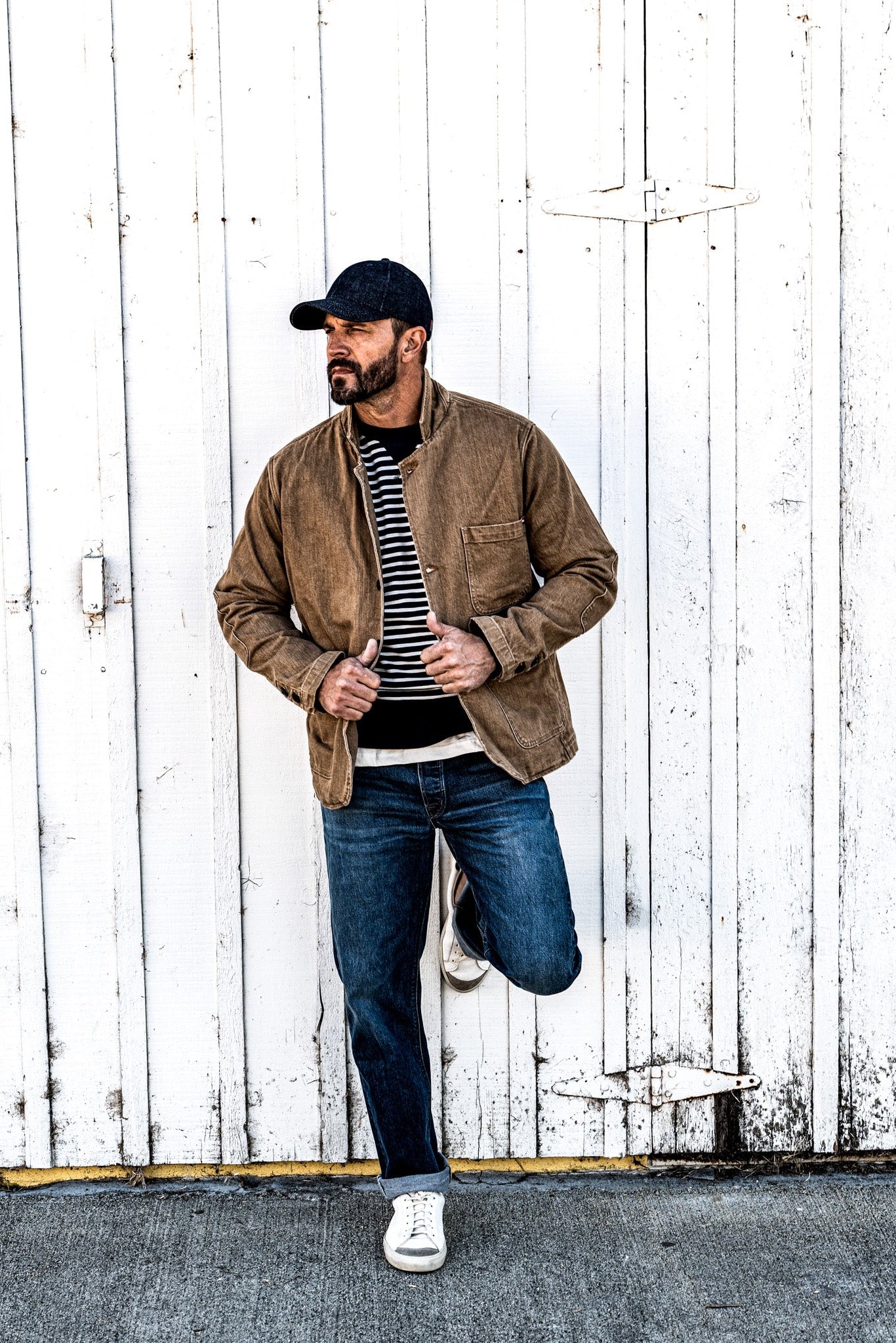 Man in a cap and a men's wheat denim chore blazer standing with hands clasped, one leg raised against a wall