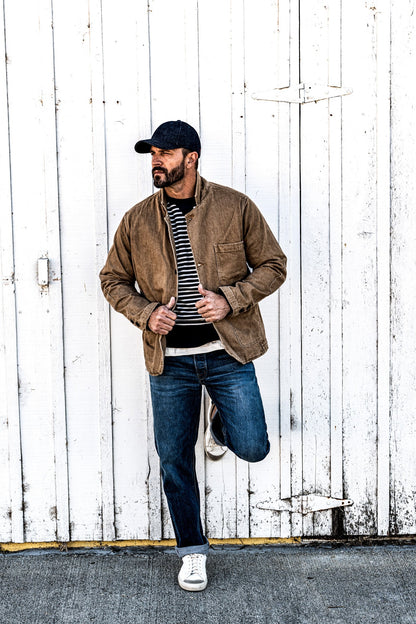 Man in a cap and a men's wheat denim chore blazer standing with hands clasped, one leg raised against a wall