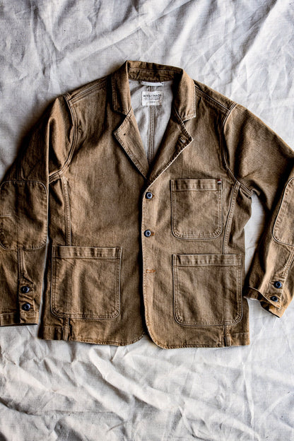 Flat lay of a men's wheat denim chore blazer with patch pockets and button-front closure on a white sheet