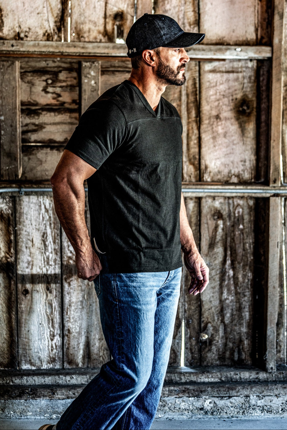 Side profile of a man in a cap wearing a men's basalt V-neck cotton jersey top and blue jeans in a barn