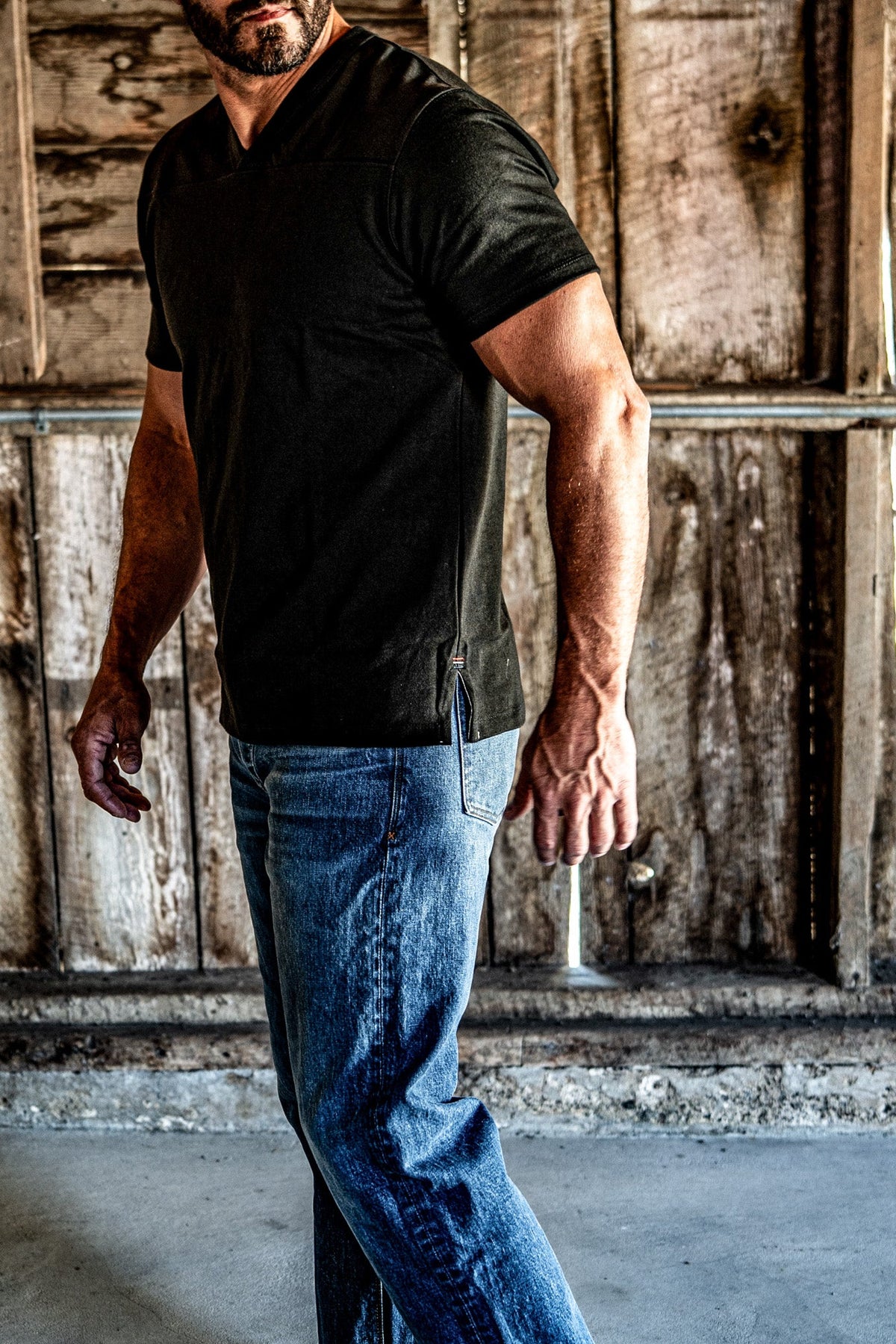 Side view of a man wearing a men's basalt V-neck cotton jersey top and blue jeans in a wooden barn