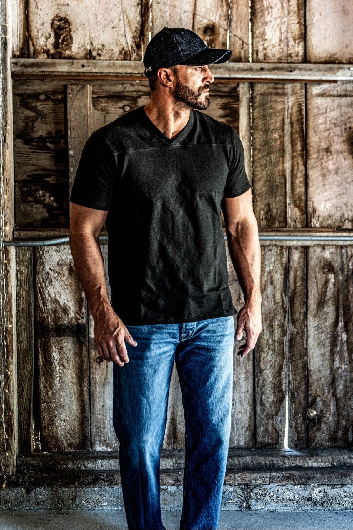 Man wearing a dark cap and a men's basalt V-neck cotton jersey top with blue jeans, standing in a rustic wooden barn