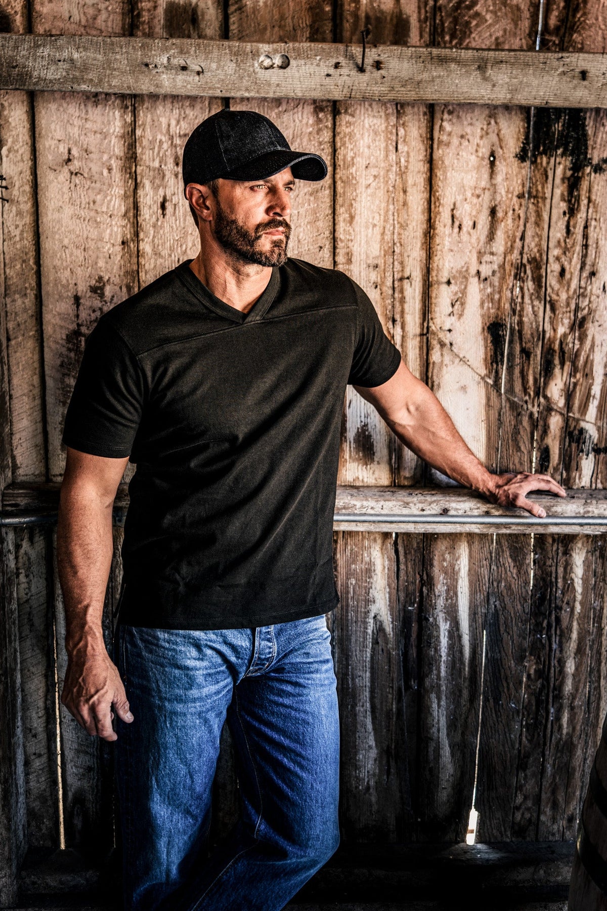 Man leaning against a wooden fence wearing a men's basalt V-neck cotton jersey top and jeans