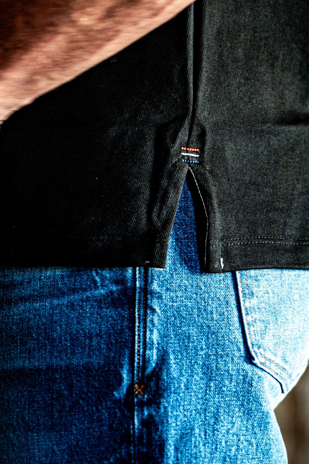 Close-up of the side hem and vent detail on a men's basalt cotton jersey top worn over blue denim jeans