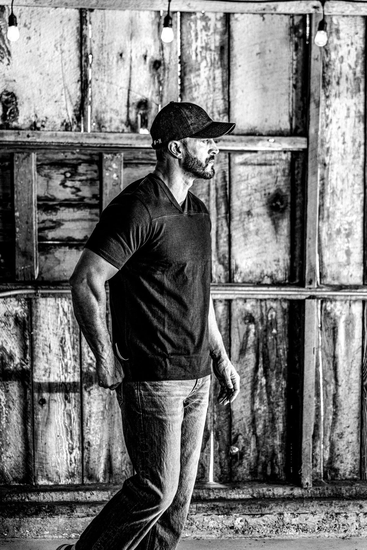 Black and white photo of a man in a men's V-neck cotton jersey top and jeans under string lights in a barn