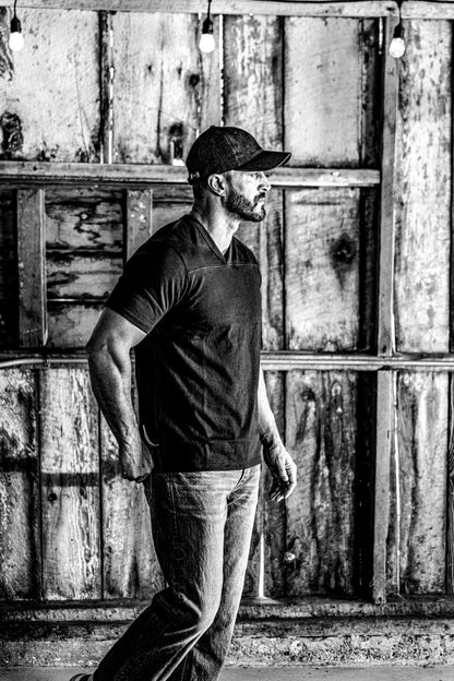 Black and white photo of a man in a men's V-neck cotton jersey top and jeans under string lights in a barn
