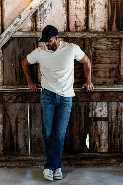Man wearing a natural heather V-neck cotton jersey top and blue denim jeans, leaning against a railing inside a rustic wooden barn