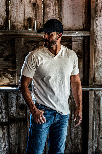Man in a baseball cap wearing a natural heather V-neck cotton jersey top and jeans beside wooden shelving in a barn