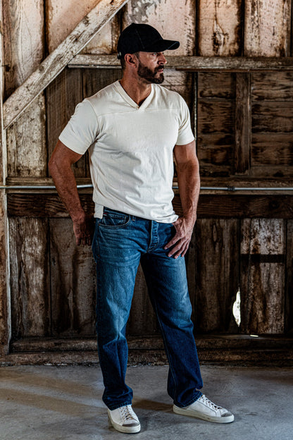 Full-body shot of a man in a natural heather V-neck cotton jersey top, jeans, and white sneakers standing in a wooden barn