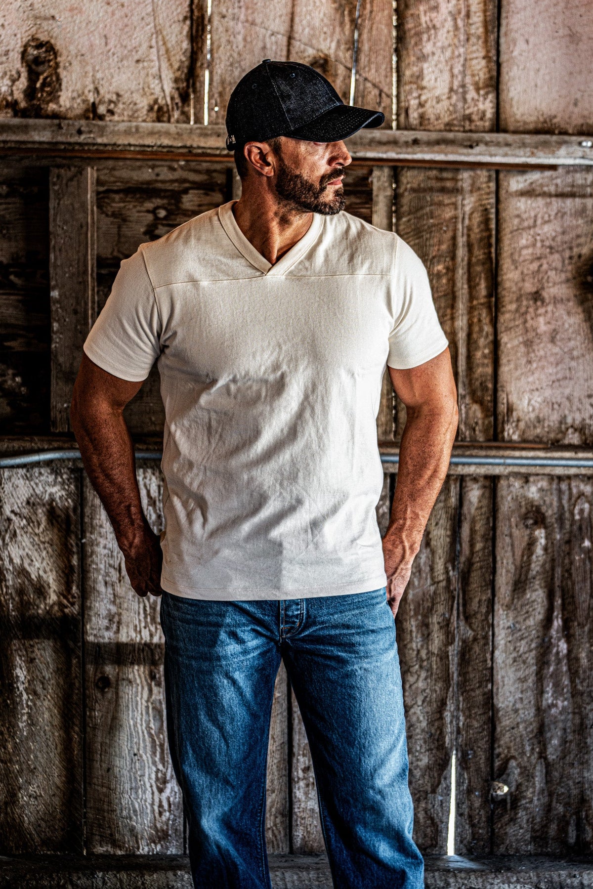 Man wearing a dark cap and a natural heather V-neck cotton jersey top with blue jeans, standing in a rustic wooden barn