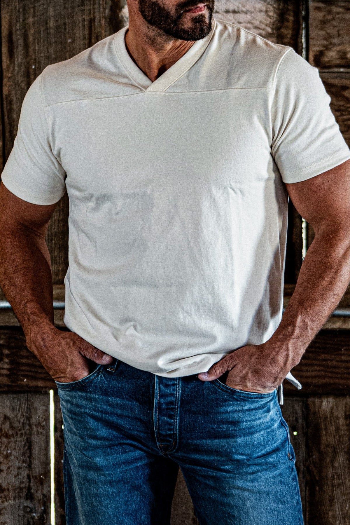 Close-up of a man's chest and shoulders in a natural heather V-neck cotton jersey top, hands tucked into denim jean pockets