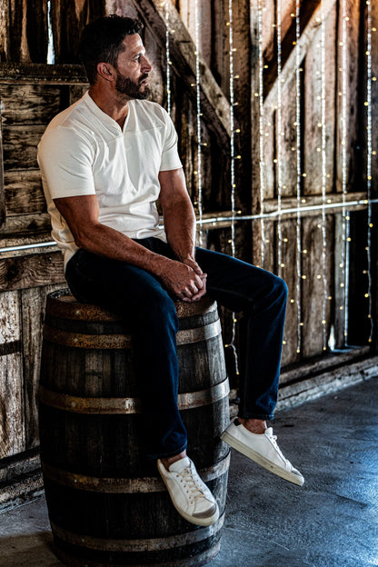 Man sitting on a wooden barrel wearing a natural heather V-neck cotton jersey top and dark jeans in a string-light-lit barn