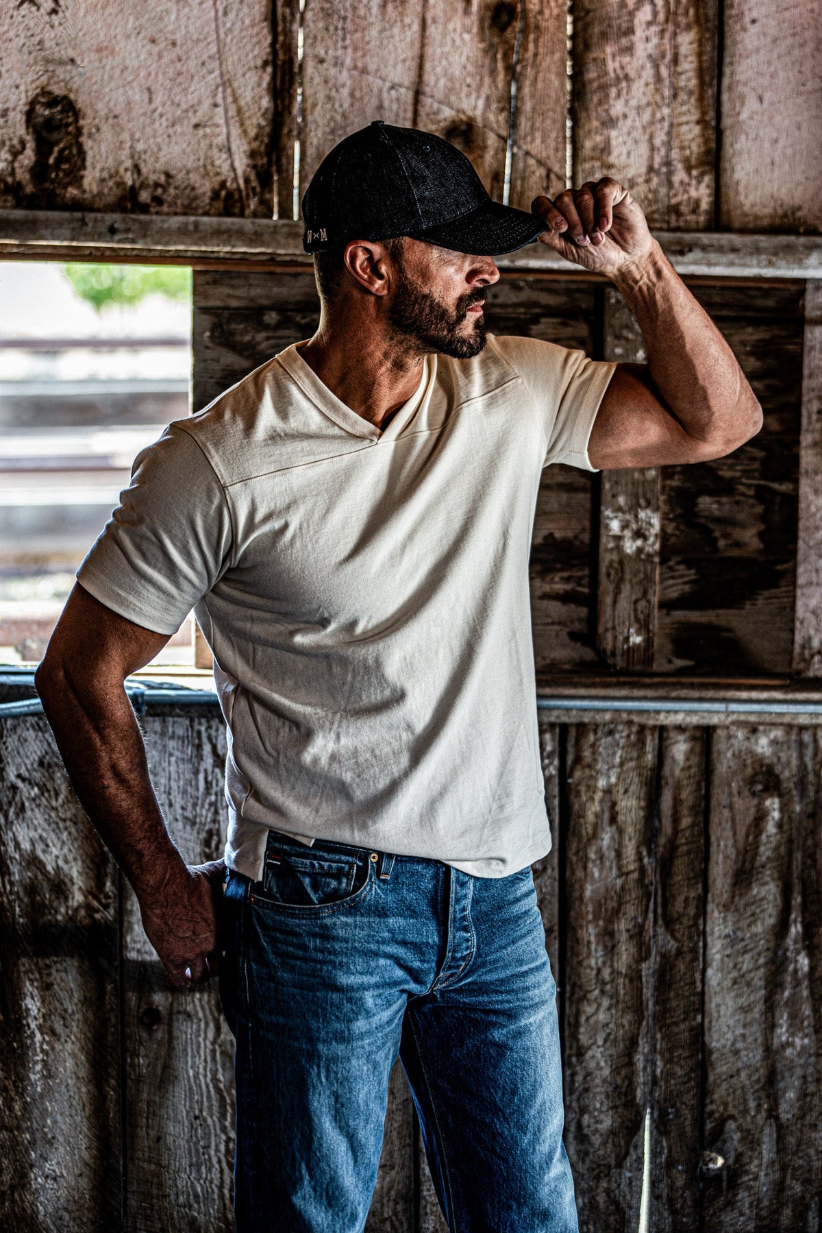 Man adjusting his cap while wearing a natural heather V-neck cotton jersey top and jeans near a barn window